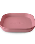 A stack of two pink, square silicone baby plates with rounded edges. "mushie" is embossed in the center. These BPA-free Square Dinnerware Plates by mushie feature a minimalist design and smooth finish.