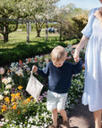 A young boy carries a mushie Water Resistant Wet Bag as he walks hand-in-hand with a woman in a light blue dress through a blooming garden on a sunny day.