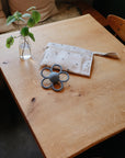 A small wooden table displays a glass jar with a leafy branch, a mushie Water Resistant Wet Bag in white with a subtle pattern for diaper storage, and a gray flower-shaped teething toy in a cozy indoor setting.