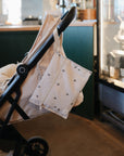 A light beige stroller is parked indoors with a cream-colored mushie Water Resistant Wet Bag featuring small black prints hanging from the handle for easy storage. Nearby, a refrigerated display case holds various products.