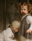 A young girl with curly hair holds a FRIGG Natural Rubber 6-Pack pacifier, standing beside a blond child who looks down. Both wear neutral-toned outfits in front of a wooden background.