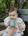 Baby on bench holding Mushie Silicone Training Cup + Straw, in patterned coat and pale leggings.