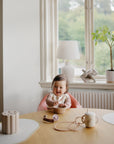 Baby in high chair with mushie Linen Pacifier Clip, eating at a table in a bright room.
