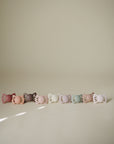 Eight FRIGG Lucky pacifiers, pastel-colored, are lined up against a beige background.
