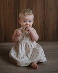 A baby in a beige dress smiles on a wooden floor, holding the mushie Pretzel Teether.