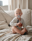 A smiling baby with a pacifier sits on a cozy bed, holding the mushie Vehicles Mold Free Bath Play Set. Sunlight filters through a window, large cushions are behind the baby, creating a warm and comfortable atmosphere.