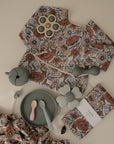 Flat lay of Mushie baby feeding items and clothing with a floral pattern, including a bib, utensils, silicone plates, flower teether, matching napkin, and the Splat Mat—a water-resistant mat in soft, muted colors.