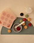 The Mushie Baby Food Freezer Tray, a sippy cup, and a bowl with spoon rest on a green placemat—ideal for baby food storage—alongside colorful round snacks. The tray features BPA-free silicone and convenient round molds.