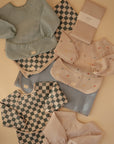 A flat lay on a beige surface featuring Mushie's Splat Mat, pastel and checkered children’s bibs and smocks, and neatly folded fabric pieces.