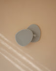 A mushie Cradle Cap Brush with gray silicone bristles and a textured oval surface is mounted on a beige wall as sunlight streams in from the left, creating soft, parallel shadows.