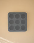 A Mushie Baby Food Freezer Tray, dark gray and square with nine round cavities, is centered on a beige surface. Soft light and shadows highlight its BPA-free silicone construction for safe baby food storage.