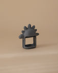 The mushie No-Drop Dino Teether (Tradewinds) is a cute gray silicone teether shaped like a stegosaurus, shown upright on a beige surface with a matching background—perfect as an adorable baby teething toy.