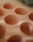 A close-up of the Mushie Baby Food Freezer Tray, a pink BPA-free silicone mold featuring round cups with volume markings in ounces and milliliters, ideal for storing homemade baby food.