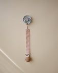 A blue pacifier hangs from a mushie Linen Pacifier Clip (2-Pack) with a wooden clasp, shown against a plain beige wall.
