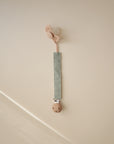 A mushie Linen Pacifier Clip 2-Pack, featuring sage green fabric straps, wooden clasps, and silicone butterfly-shaped pacifier holders, rests on a beige surface.