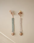 Two mushie Linen Pacifier Clip 2-Packs with wood ends hang on a beige wall.