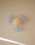 The mushie Suction Spinner Toy features a beige sun and three blue clouds, all made from food-grade silicone, mounted on a beige wall and softly lit in the lower left corner.