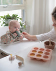 A baby sits in a high chair, drinking from a cup as an adult holds their arm. On the white table are a bowl, silicone mold tray, pacifier, and the mushie No-Drop Cactus Teether (Dried Thyme), all bathed in bright natural light.