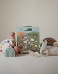 A selection of pastel mushie baby toys is displayed on a white background, featuring stacking rings, a rainbow stacker, animal-themed teethers, puffy stickers, the mushie Reusable Sticker Set, and a soft cloth.