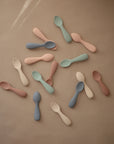 A selection of mushie Silicone Toddler Starter Spoons in pastel pink, green, blue, beige, and brown is arranged on a plain brown background—ideal for independent feeding.