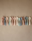 A row of mushie Silicone Toddler Starter Spoons, designed for independent feeding, displayed side by side on a plain tan surface in soft pastel shades.