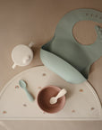 The mushie Silicone Toddler Starter Spoons are part of a pastel baby feeding set featuring a green silicone bib, sippy cup, bowl, and rainbow placemat—ideal for encouraging independent feeding on beige surfaces.