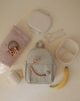 Flat lay of baby items on a light surface: a soft pink blanket with a beaded teether, a beige backpack with the mushie Silicone Pacifier Clip | Luna, a divided lunch container, utensils, lidded cup, and ripe banana.