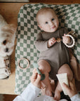 Baby lies on a green-checkered mat; use mushie Baby Cream to soothe dry skin.
