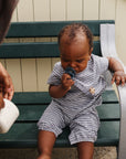 Baby in checkered outfit holds FRIGG Moon Natural Rubber Pacifier on green bench with adult.