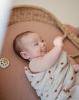 A baby smiles in a woven bassinet wrapped in a light-patterned blanket, while a FRIGG Andersen Fairytale Natural Rubber Pacifier 2-Pack rests nearby on the mattress.