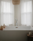 A modern white bathtub with a wooden tray holds mushie's Fragrance Free Baby Body Lotion, set in front of sheer white curtains and lit by natural light, with a woven pendant overhead and a small table beside the tub.