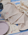 A pastel picnic setup includes mushie's Dinnerware Cutlery Set, a lavender plate, gray cup, cream smiley face napkins, all on a blue and white striped blanket with a dotted cloth nearby.