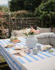A picnic table with a blue and white striped tablecloth is set outdoors with a vase of flowers, plates, fruit, and the Mushie Bluey x Mushie Silicone Training Cup + Straw. Cushioned seating and greenery in the background create a cozy garden vibe.