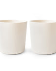 Two mushie Dinnerware Cups sit side by side on a white background. These non-toxic, plain white ceramic cups are cylindrical with smooth surfaces, lacking handles or decorations.