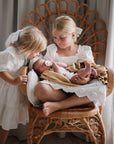 Two girls admire a newborn with a FRIGG Daisy Natural Rubber Pacifier in a cozy wicker chair.
