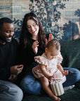A smiling man and woman sit on a couch with a toddler girl in a pink dress, who is playing with the mushie Coin & Tube Sorting Set. The adults look happy and engaged, and there’s a decorative tapestry in the background.