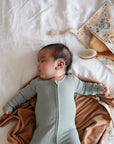 A baby in a light green onesie lies on a bed with a brown blanket, next to a mushie Water Resistant Wet Bag holding essentials like a hairbrush, pacifier, and other baby care items.