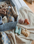 A neatly organized box with folded baby clothes sits on a bed beside a beige towel featuring the mushie Nature Mold Free Bath Play Set—mold-free, food-grade silicone bath toys shaped like binoculars, a squirrel, dinosaur, and acorn.