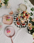 Mushie Round Dinnerware Plates, forks, pastries, macarons, and flowers on a white tablecloth.