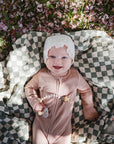 A smiling baby in a pink outfit lies on a mushie Organic Cotton Muslin Swaddle Blanket atop a black-and-white checkered surface outdoors, surrounded by pink petals and green grass.