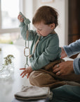 A toddler uses a FRIGG Butterfly Anatomical Silicone Pacifier (0-6 Months) while sitting at a table, holding a toy. An adult supports the child in a cozy, softly lit room decorated with vases of flowers and folded clothes.