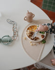 A child in a checkered outfit enjoys pasta, corn, and broccoli from a divided plate at a white table, using the mushie Dinnerware Fork and Spoon Set; a cup and green silicone toy are nearby.