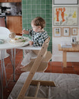 A young child eats from a bowl in a high chair at the dining table, with a Mushie Splat Mat under the chair protecting the floor. The kitchen features green tiles and framed artwork, creating a cozy, homey feel.