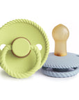 The Mushie FRIGG Rope Natural Rubber Pacifier 2-Pack includes two baby essentials: a light green pacifier with a handle standing upright and a light blue one on its side, both featuring rope-like edges and natural rubber latex nipples.