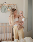 A woman with long blonde hair in a white top and yellow pants holds a baby in beige in a softly lit, minimal nursery with a wooden crib featuring the mushie Stretchy MINI Crib Sheet and neutral decor.