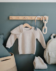 A small beige knit sweater hangs on a wooden peg rack against a sage green wall, with a folded matching garment, beige backpack, basket, and FRIGG Butterfly Anatomical Silicone Pacifier 2-Pack (0-6 Months) nearby. Large chain links hang from the rack.