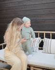 A woman with long blonde hair sits on a bamboo bench against a dark wood wall, holding a toddler in green knit clothes using the mushie Silicone Pacifier Clip | Luna. The bench is topped with patterned cushions.