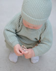 A baby in a light green knit outfit and hat squats on a light surface, playing with a FRIGG Lucky Symmetrical Silicone Pacifier (0-6 Months) in both hands. The white socks on the baby's feet are visible.