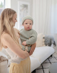 A woman with long blonde hair holds a baby in a light green knit outfit and hat, featuring the mushie Silicone Pacifier Clip | Luna, in a bright, cozy living room with white and beige decor.