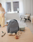A small gray backpack, baby toys, and the mushie Silicone Pacifier Clip | Luna rest on a wooden table in a bright, cozy living room. A woman in yellow pants and a white top stands blurred near the window in the background.